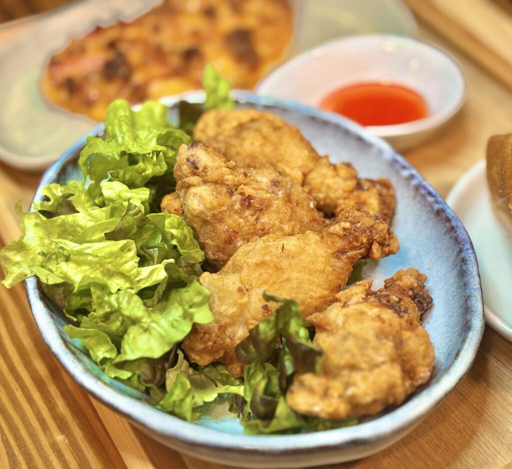 レタスを添えたフライドチキンを器に盛り、奥にスィートチリソースと鶏胸肉ピザが並ぶ食卓写真