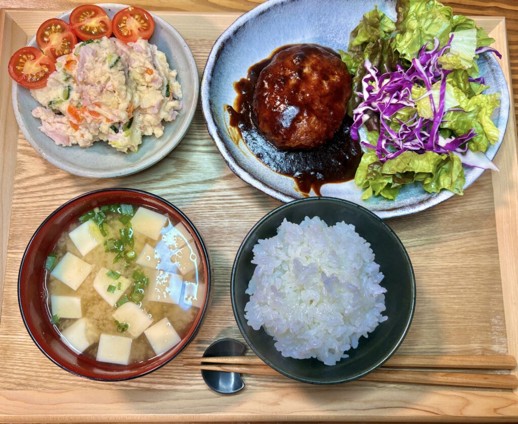 木のトレーの上に並んだ家庭料理の夕食。デミグラスソースのチーズインハンバーグ、レタスと紫キャベツのサラダ、ポテトサラダ、豆腐の味噌汁、白ごはんがバランスよく配置されている定食風の食卓。