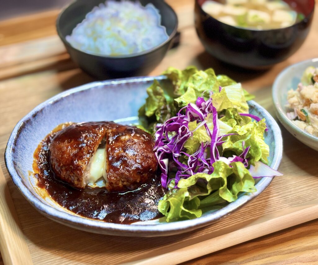 半分にカットされたチーズインハンバーグからとろりとチーズが溶け出し、濃厚なデミグラスソースがたっぷりかかったメイン料理。横にはレタスと紫キャベツのサラダを添えている。