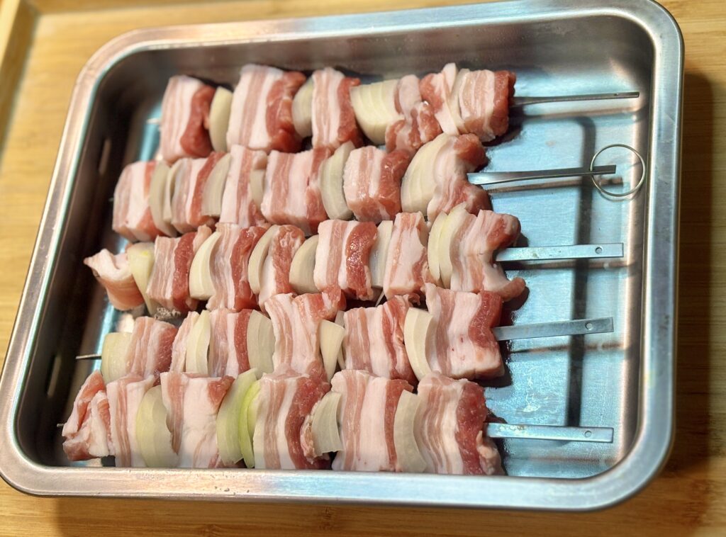 バットに並べられた豚バラと玉ねぎの串。焼く前の下ごしらえ状態で、均等に刺さった具材が整然と並んでいる様子。