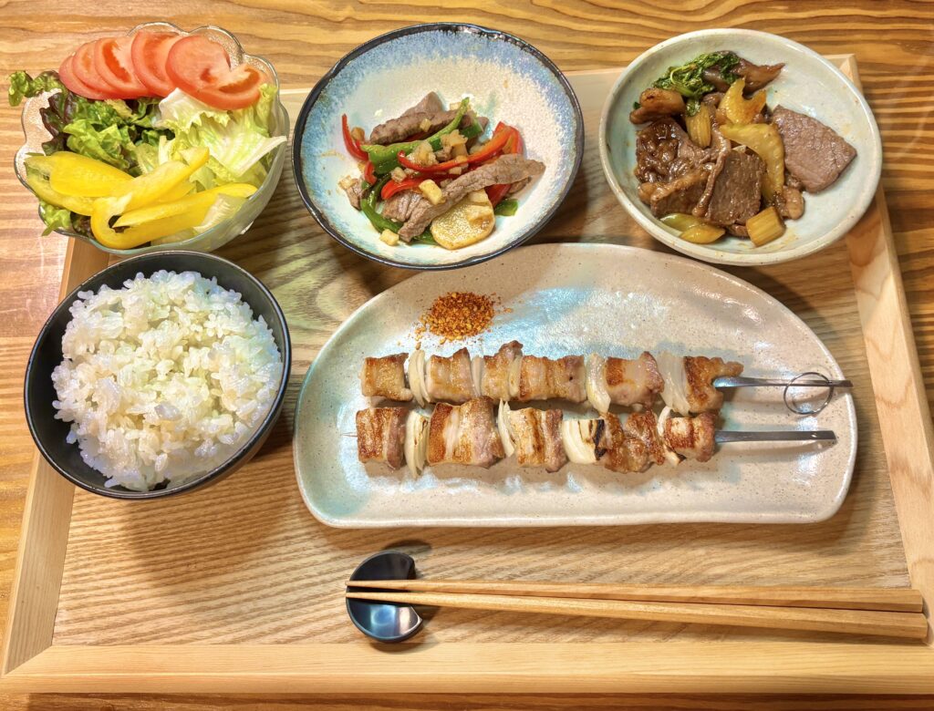 豚バラと玉ねぎの串焼きを中心に、ごはん、サラダ、牛肉のおかずが並んだ手作り定食。木製トレーに盛りつけた家庭的で温かみのある食卓風景。