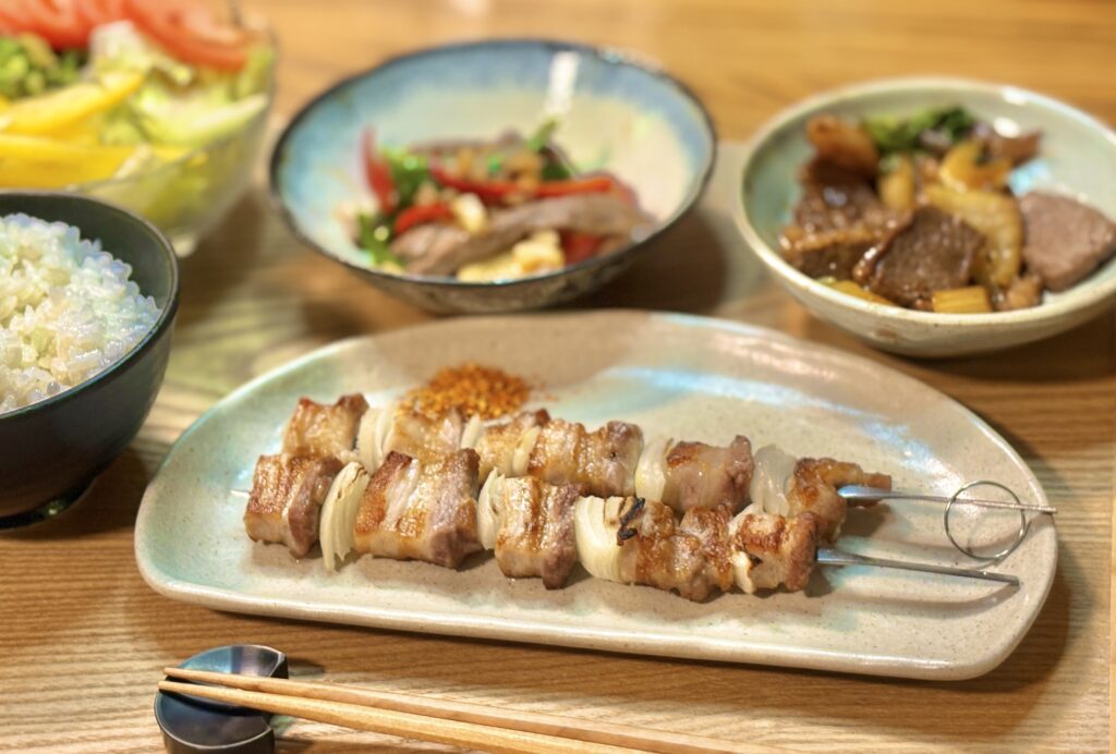 こんがり焼けた豚バラ玉ねぎ串を中心に、ごはん、サラダ、牛肉炒め、小鉢が並ぶバランスのよい和風献立。