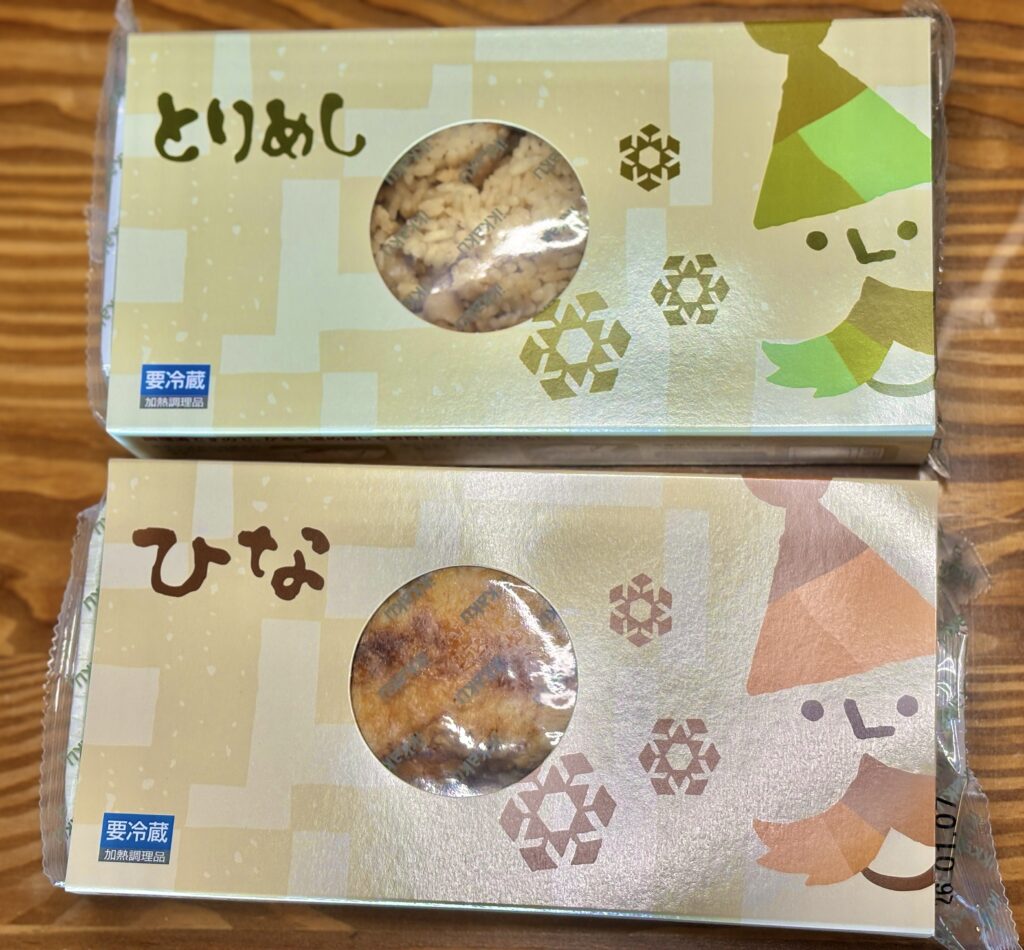 鶏のひな焼きと鶏めしのテイクアウト商品。専用パッケージに入った状態と、透明フィルムで包装された鶏のもも焼きと鶏めしが木目のテーブルの上に並んでいる。調理前の持ち帰り用商品として内容が分かる写真。