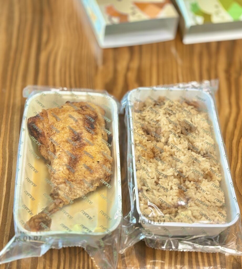 鶏のひな焼きと鶏めしのテイクアウト商品。専用パッケージに入った状態と、透明フィルムで包装された鶏のもも焼きと鶏めしが木目のテーブルの上に並んでいる。調理前の持ち帰り用商品として内容が分かる写真。