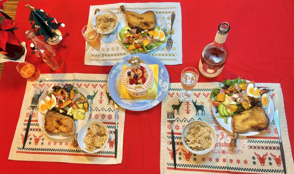 赤いテーブルクロスの上に3人分のクリスマスディナーが並ぶ食卓。ローストチキン、半熟ゆで卵と彩り野菜のサラダ、炊き込みご飯、小鉢料理がそれぞれのプレートに盛られ、中央にはホールケーキが置かれている。ロゼのスパークリングワインとグラス、クリスマスツリーの飾りがあり、華やかで温かみのある家庭の食卓風景。