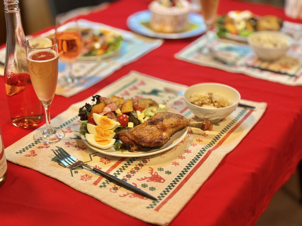 赤いテーブルクロスの食卓に並ぶクリスマスディナー。ローストチキン、半熟ゆで卵と野菜のサラダ、小鉢料理がワンプレートに盛られ、横にはロゼのスパークリングワインがグラスに注がれている。温かみのある家庭的な食卓風景