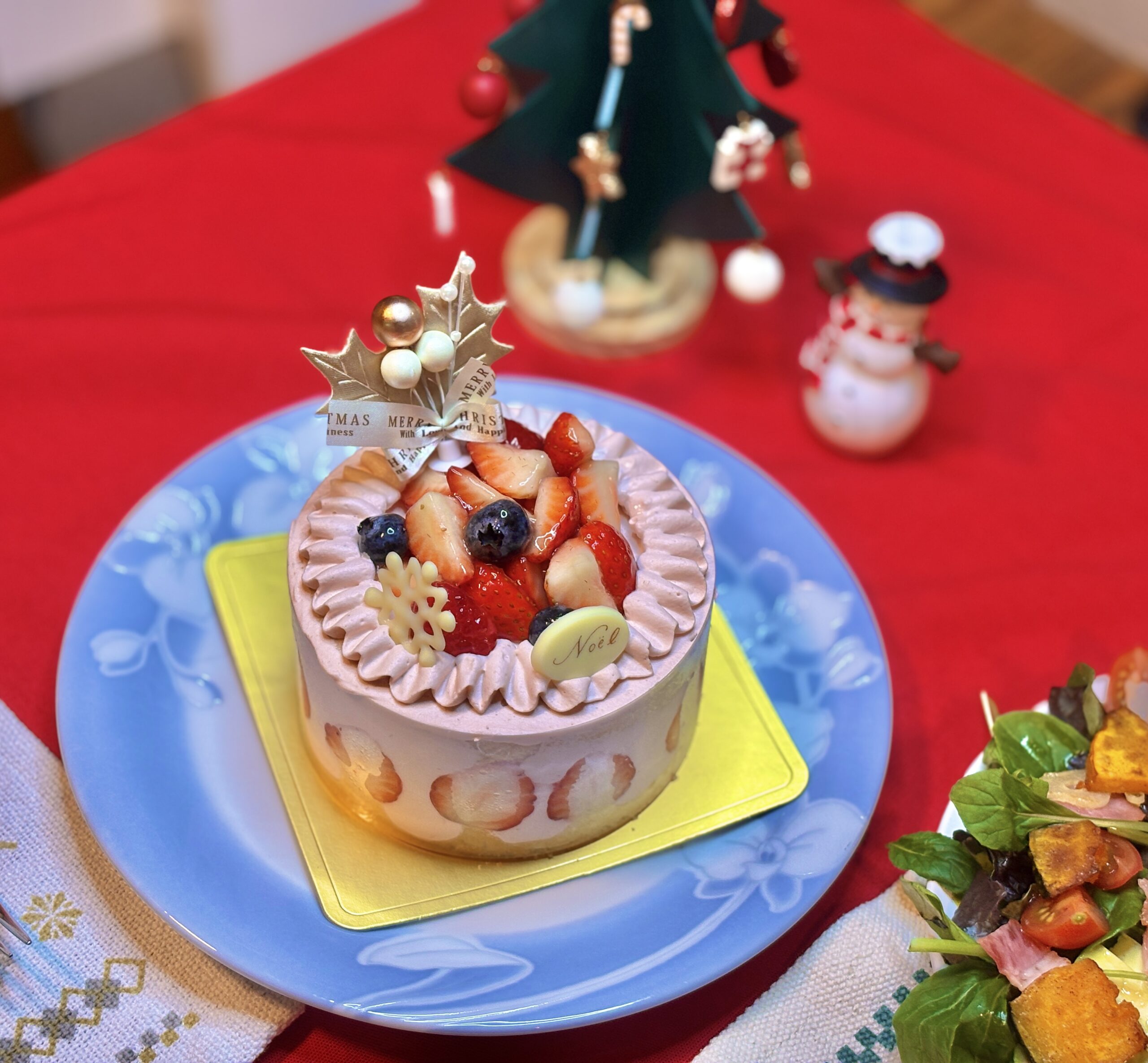 クリスマスのテーブルに並べられた苺とブルーベリーたっぷりの小さなホールケーキ。赤いテーブルクロスの上に、ツリーと雪だるまの飾りが背景に写っている。