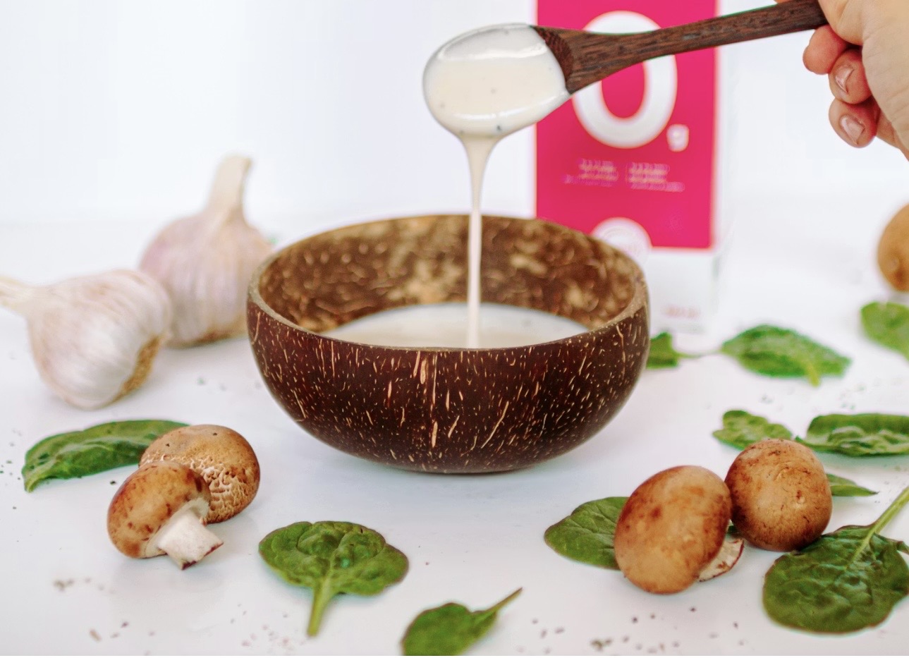 ココナッツボウルに入った白いソースを木のスプーンですくっている様子。周囲にはマッシュルームやほうれん草、にんにくが並び、ナチュラルでヘルシーな料理イメージの写真。