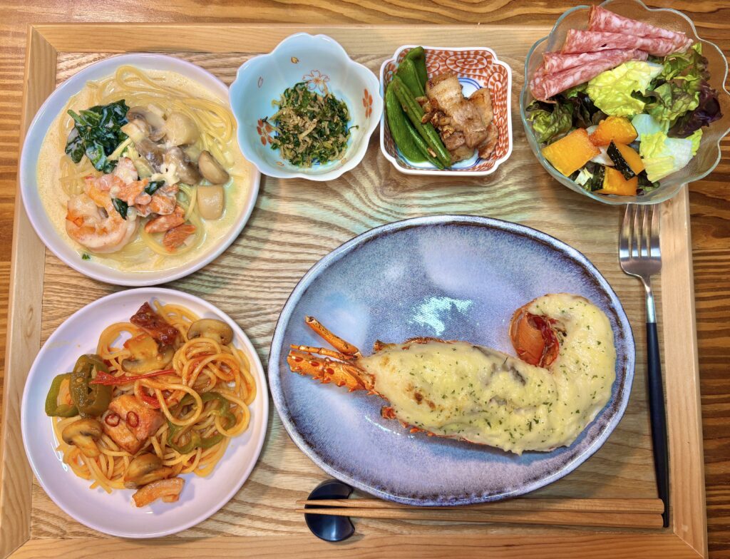 木のトレーに伊勢海老のテミドールをメインに、シーフードとほうれん草のクリームパスタ、トマトソースパスタ、小鉢2種、サラダを並べた洋風の食卓