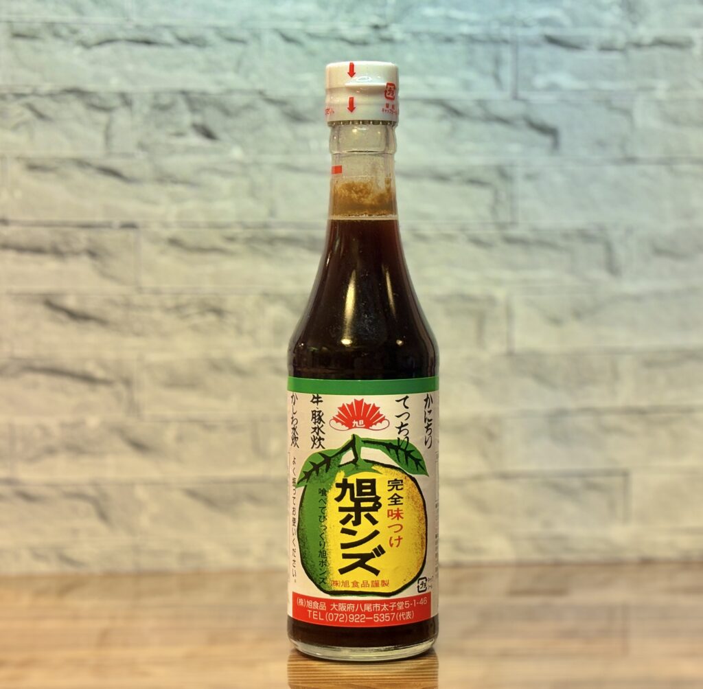 旭食品の調味料「旭ポンズ」の瓶を正面から撮影した写真。柑橘のイラストが描かれたラベルが見え、濃い色のポン酢が入った瓶が木目調の背景の前に置かれている