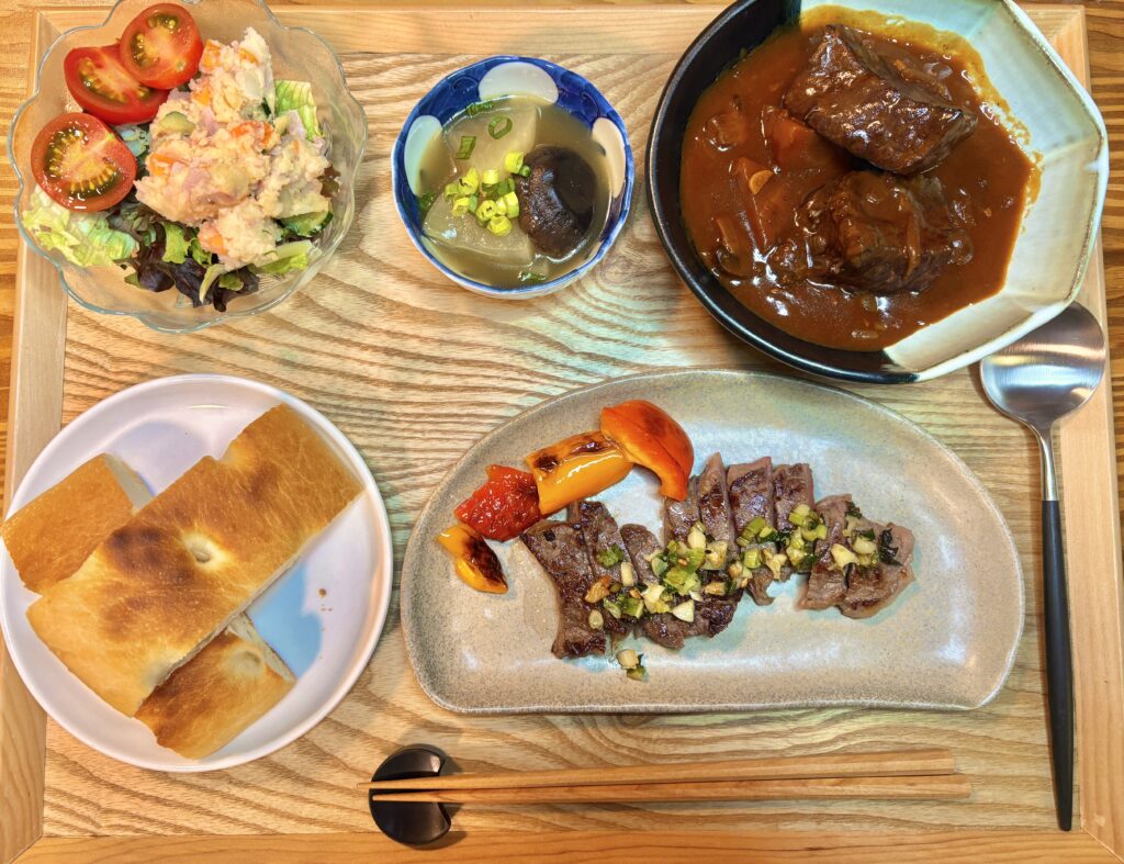 牛ステーキのガーリックネギソースと焼きパプリカ、黒毛和牛の赤ワイン煮込み、大根と椎茸の鶏がらスープ煮、ポテトサラダとフォカッチャを木製トレーに並べた、おうちビストロ風の贅沢ディナー。