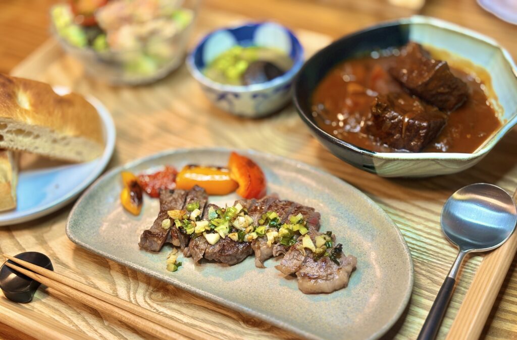 ガーリックネギソースをかけた牛ステーキを器に盛り付け、焼きパプリカを添えた一皿。木製トレーの上にフォカッチャや赤ワイン煮込みが並ぶ、おうちビストロ風ディナーの一場面。
