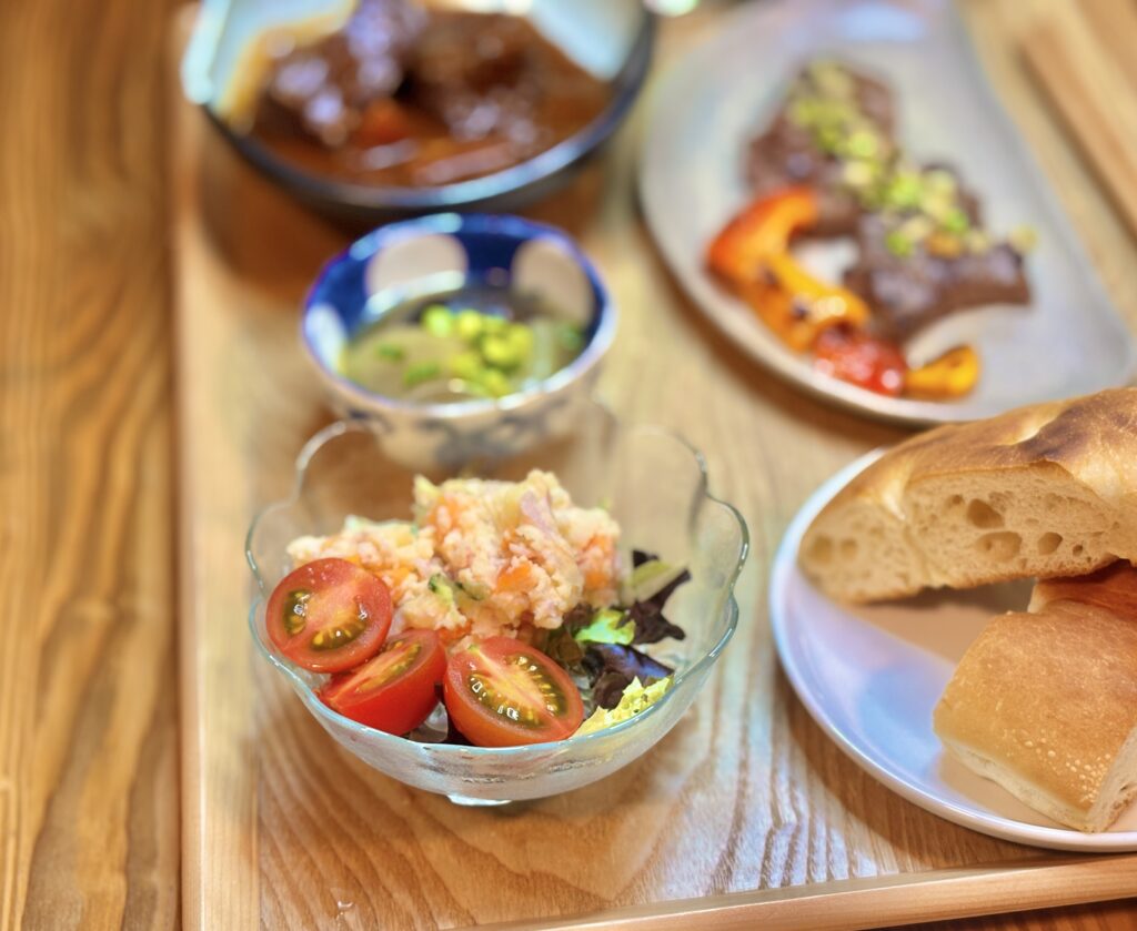 ポテトサラダとミニトマトを盛り付けたガラスの器と、横に添えたフォカッチャを木製トレーに並べた一皿。奥には大根と椎茸の鶏がらスープ煮や牛ステーキがぼんやり映る、おうちビストロ風の食卓。