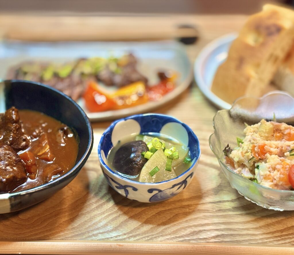 黒毛和牛の赤ワイン煮込み、大根と椎茸の鶏がらスープ煮、ポテトサラダを木製トレーに並べた副菜中心の盛り付け。奥には牛ステーキとフォカッチャが見える、おうちビストロ風ディナーの一場面。