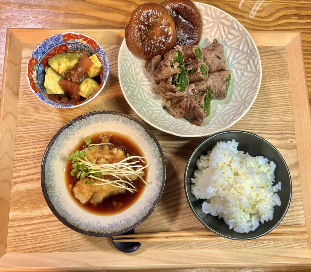 木製トレーに並んだ和風献立。水菜・春菊・ほうれん草を巻いた牛肉巻きに、おろしあさひポン酢をかけた主菜。香ばしく焼いた肉厚の椎茸。小鉢にはアボカドとマグロの角切りをわさび醤油で和えた一品。ご飯と副菜を添えた、栄養バランスの良い家庭料理の食卓風景