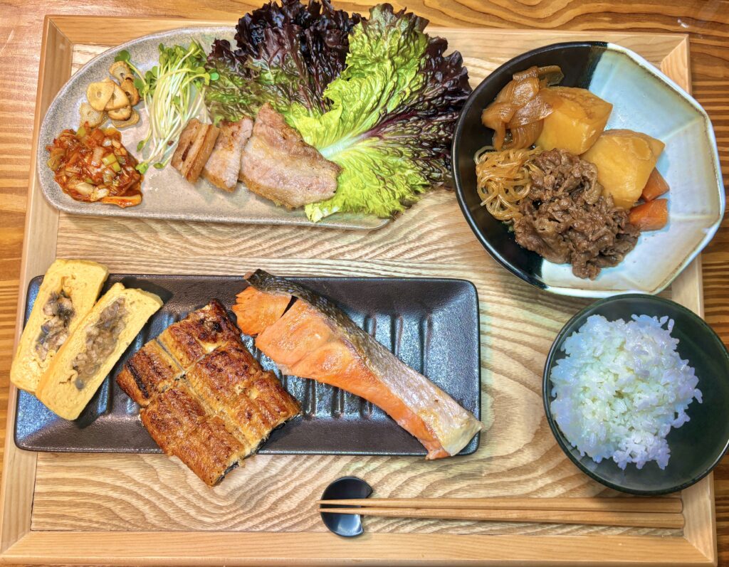 木のトレーに並んだおうちごはんの献立。阪神百貨店の魚伊の鰻巻きと鰻の蒲焼き、塩しゃけ、肉じゃが、サムギョプサル、白ごはんが盛り付けられている。