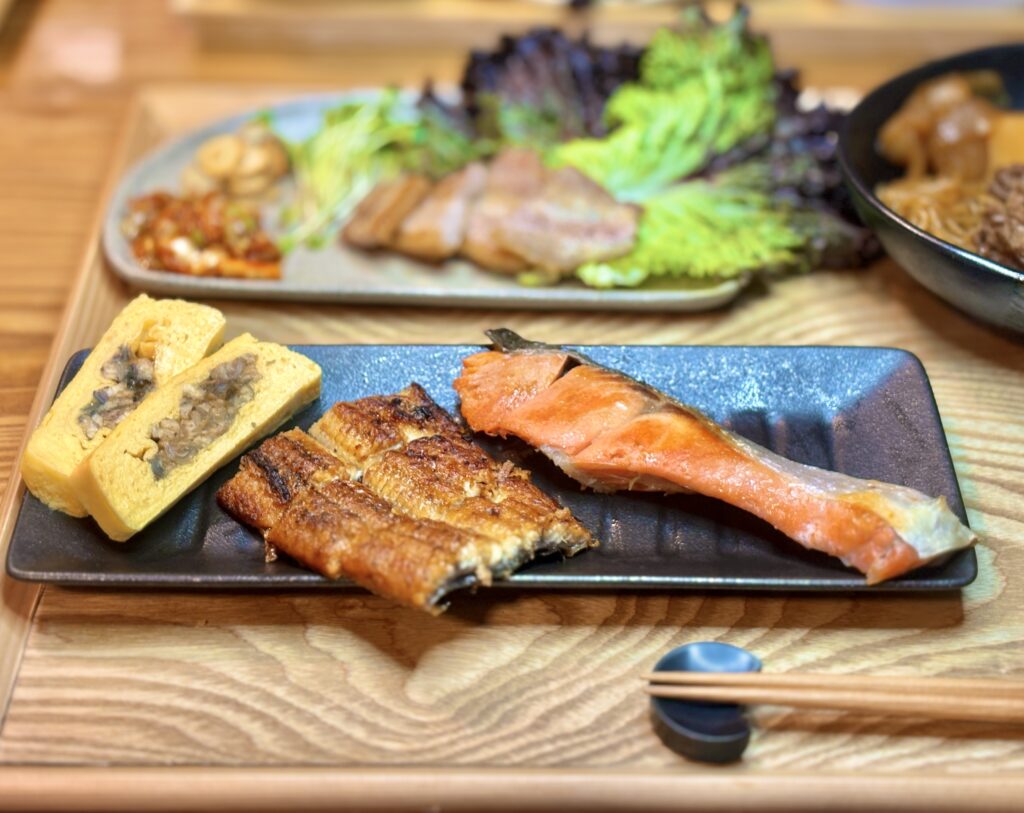 黒い長皿に盛り付けた阪神百貨店・魚伊の鰻の蒲焼きと鰻巻き、塩しゃけ。奥にはサムギョプサルと副菜、肉じゃがが並ぶおうちごはんの食卓。