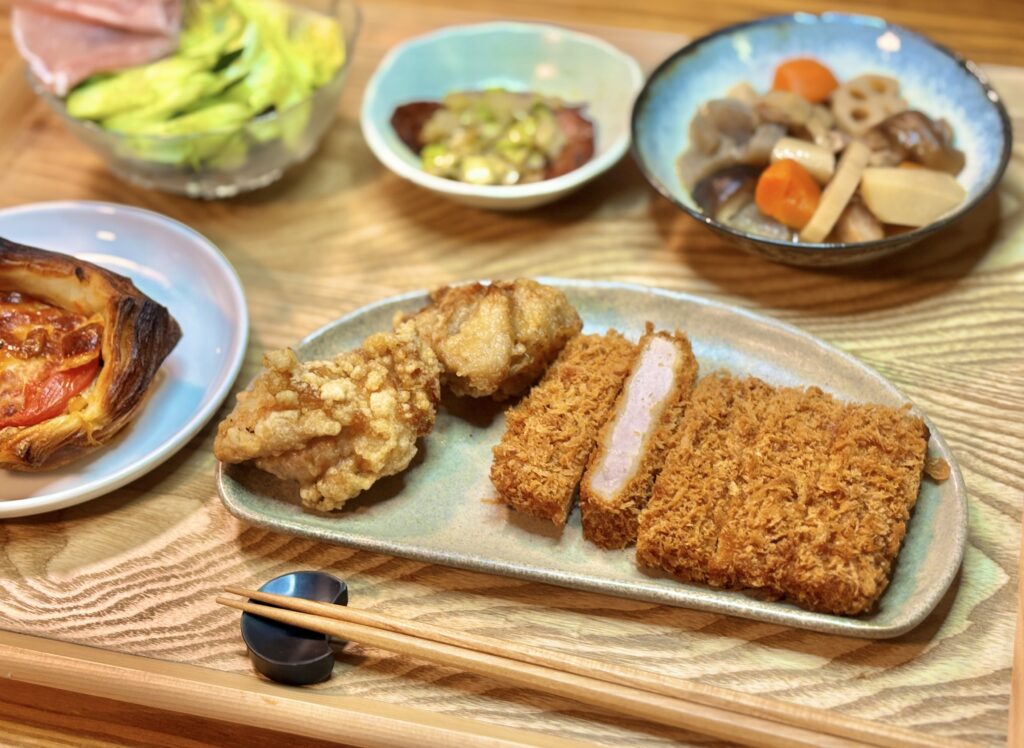 木目のテーブルに並んだ和洋折衷の食卓。手前の皿にはミュンヘン風の唐揚げと、断面が見えるまい泉のヒレカツ。周囲に博多の郷土料理がめ煮、ネギをのせたチャーシュー、生ハムサラダ、トマトとツナのパンが配置された食事風景