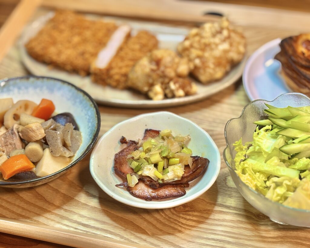 木目のテーブルに並んだ家庭の食卓。奥にまい泉のヒレカツとミュンヘン風の唐揚げ、手前には博多の郷土料理がめ煮、ネギをのせたチャーシュー、生ハムとレタスのサラダ、小皿の惣菜が並ぶ料理写真