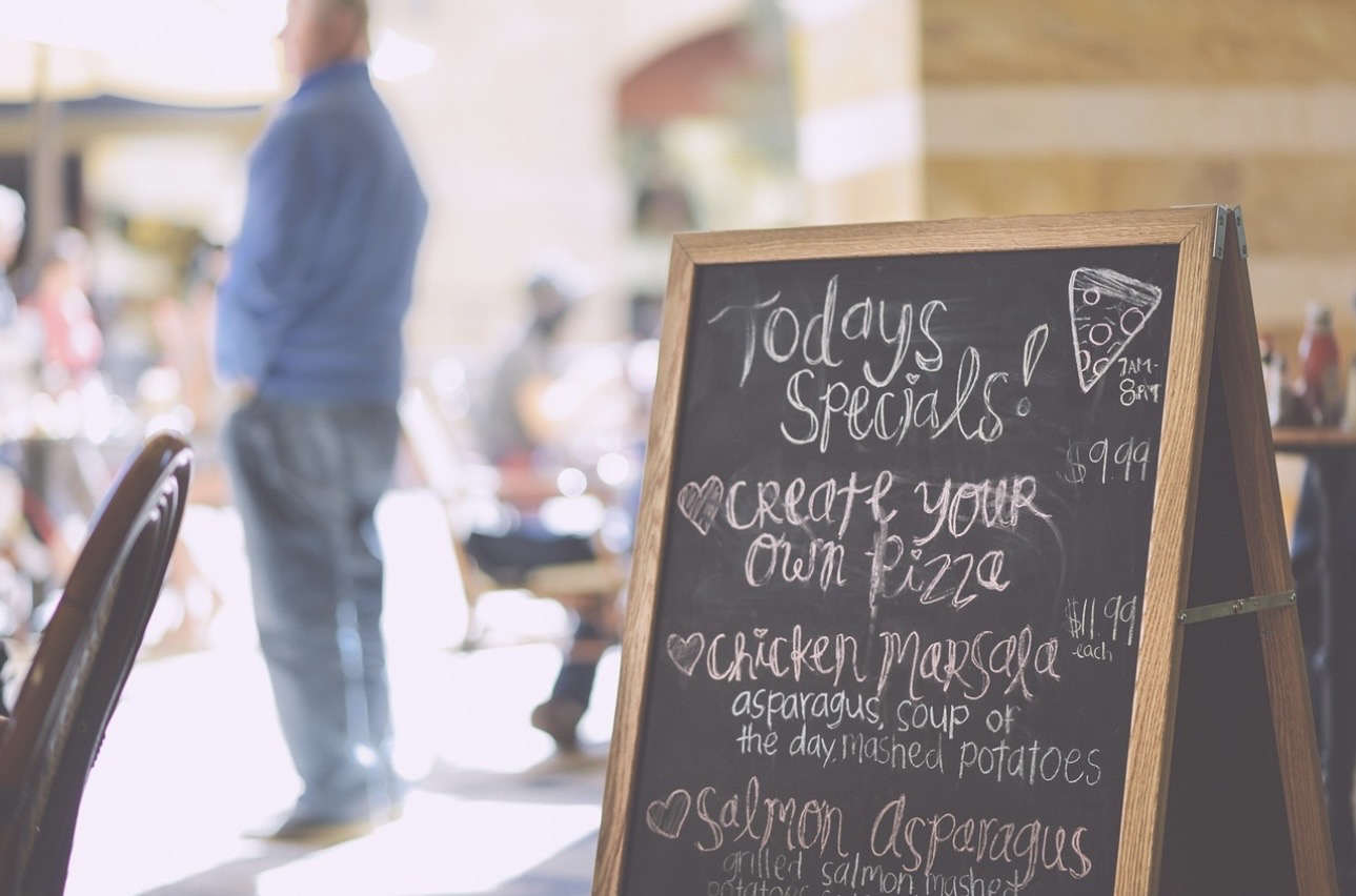 カフェ店内に置かれた黒板の本日のおすすめメニュー。オリジナルピザ、チキンマルサラ、サーモンとアスパラガスがチョーク文字で書かれ、奥には人が行き交う明るいカフェの雰囲気が広がっている。