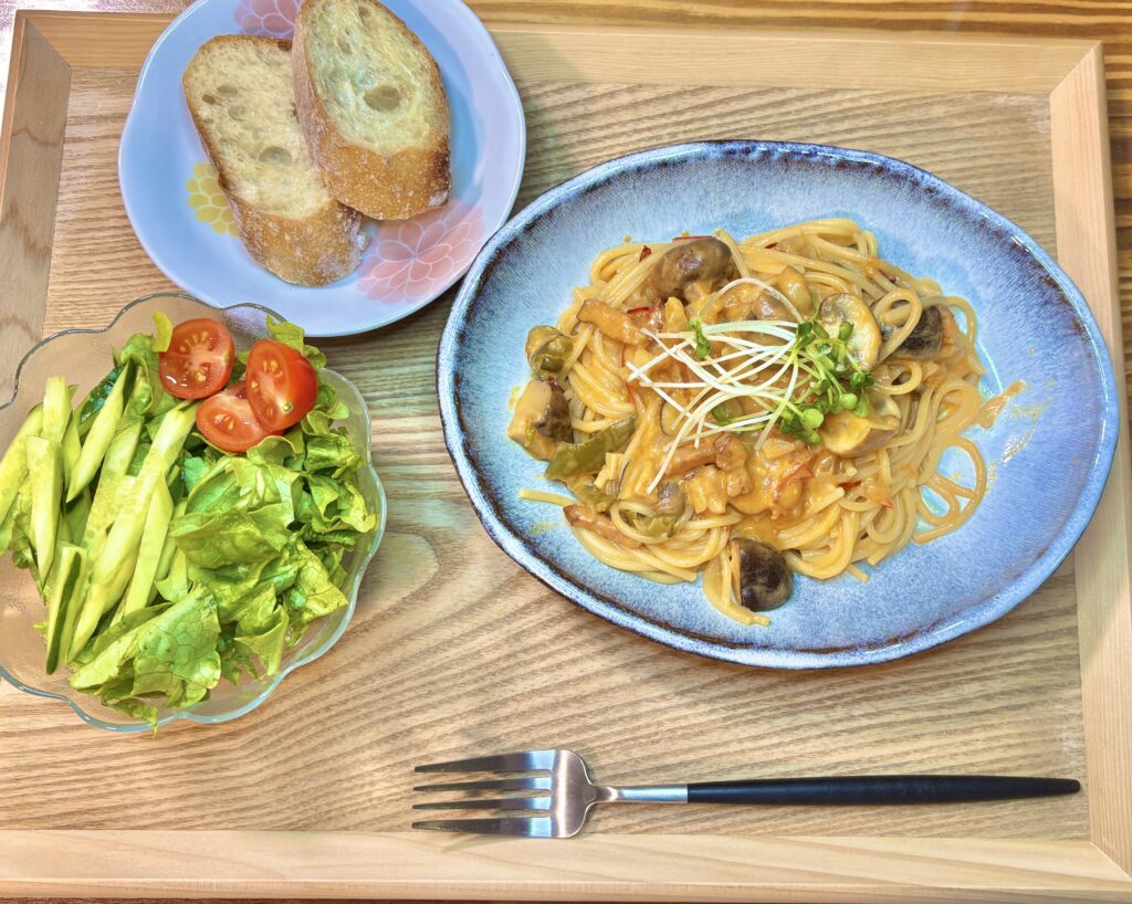 木製トレーの上に盛り付けられた夜ごはん。椎茸とマッシュルーム、ベーコンのトマトクリームソースパスタを中心に、バゲットとレタス・きゅうり・ミニトマトの野菜サラダが並ぶ家庭的な食卓。