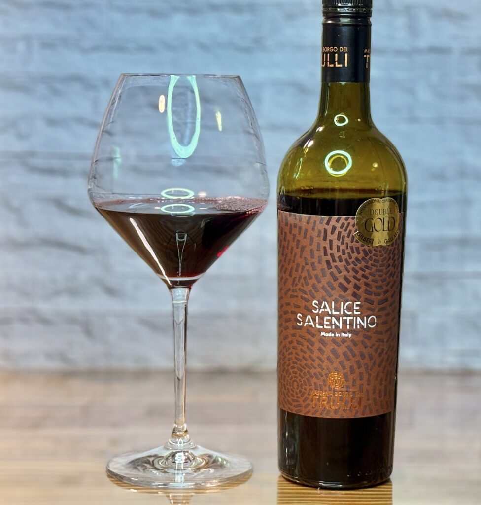 赤ワインが注がれたワイングラスと、イタリア産赤ワイン「サリーチェ・サレンティーノ」のボトル。落ち着いた背景の前に並べられた、夜の食卓を彩る一杯。