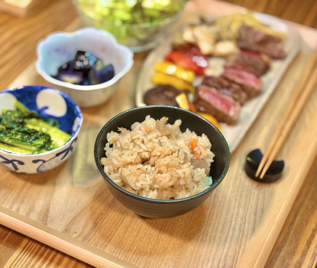 アンガス牛ステーキ献立の主食、昨日の残りを活用した具だくさん鶏飯(炊き込みご飯)。手前の黒いお茶碗に盛られた鶏とごぼうの旨味たっぷりのご飯と、奥に並ぶメインのお肉料理。