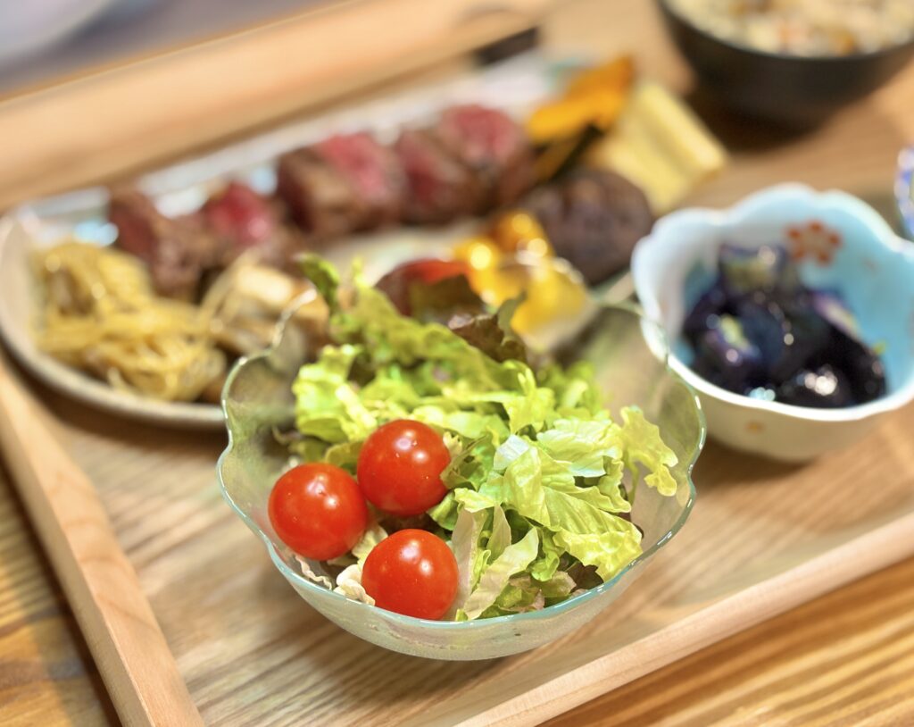 アンガス牛ステーキ献立の付け合わせ、レタスとミニトマトのフレッシュサラダ。手前のガラスボウルに盛り付けられた鮮やかな野菜と、奥に広がるメインディッシュの食卓風景。