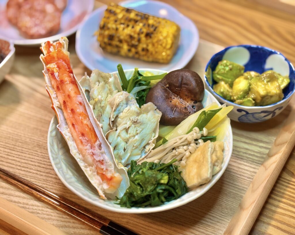 旦那キッチンの豪華な晩御飯。器からはみ出すほど大きなタラバガニの脚と、屋台の焼きとうもろこし、アボカドの副菜が並ぶ食卓
