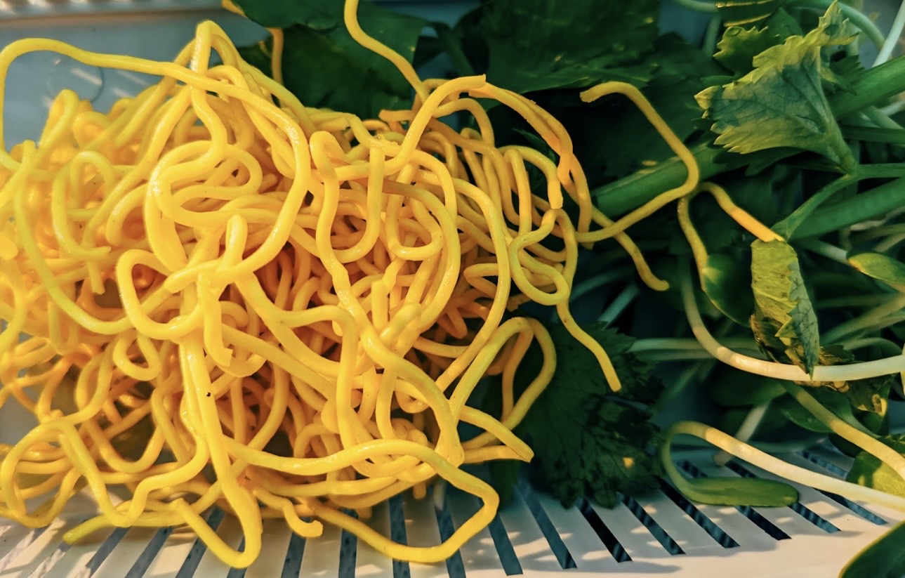 黄色い中華麺と、三つ葉や水菜などの青菜を並べた料理写真