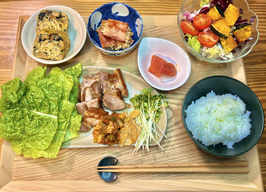 木製トレイに並んだ豪華な定食。メインの豚バラ焼きとサンチュ、白米、かぼちゃのサラダ、ひじきと豆入りの卵焼き、えのきのベーコン巻き、明太子が彩りよく配置されている。