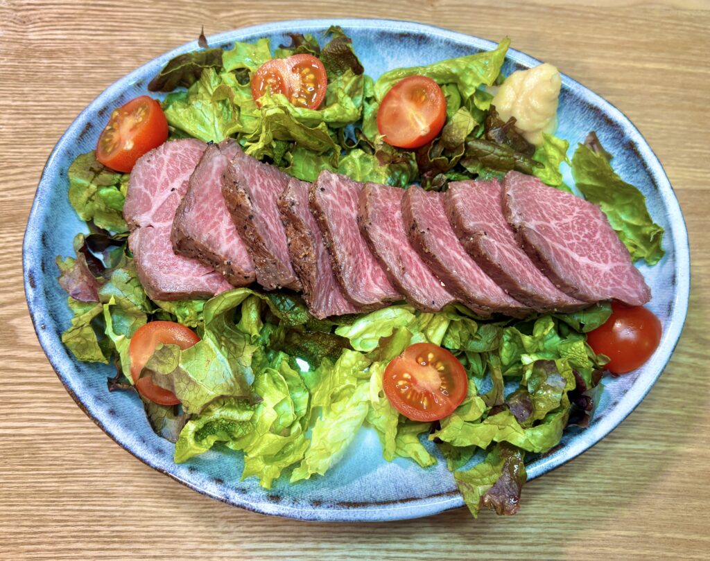 青い陶器の皿に贅沢に盛り付けられた、サシの美しい黒毛和牛のローストビーフ。新鮮なレタスとミニトマト、そしてツンと辛いホースラディッシュが添えられた一皿。