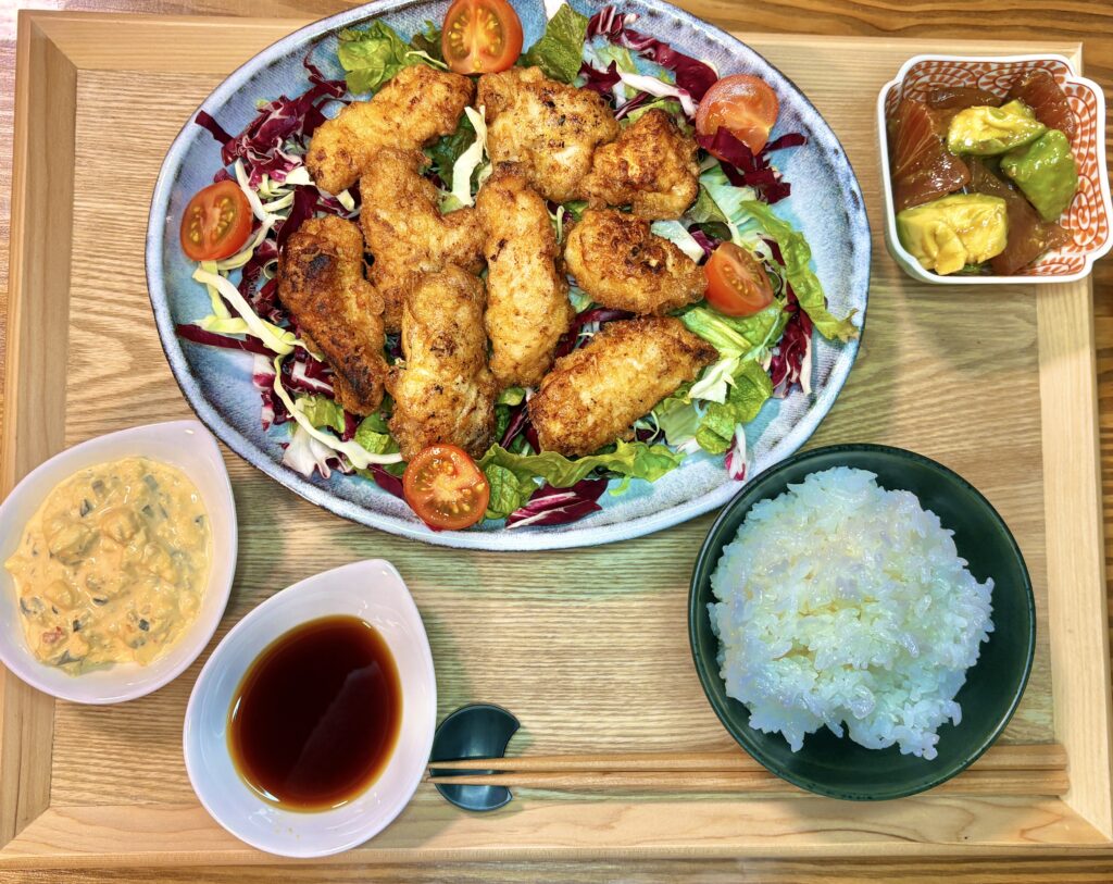 木製トレーに乗った豪華な夕食のセット。メインの一口サイズにカットされたチキン南蛮と色鮮やかな野菜サラダ、マグロとアボカドのワサビ和えの小鉢、ふっくらと炊き上がった白米、そしてたっぷりのタルタルソースと甘酢ダレが並んでいる様子。
