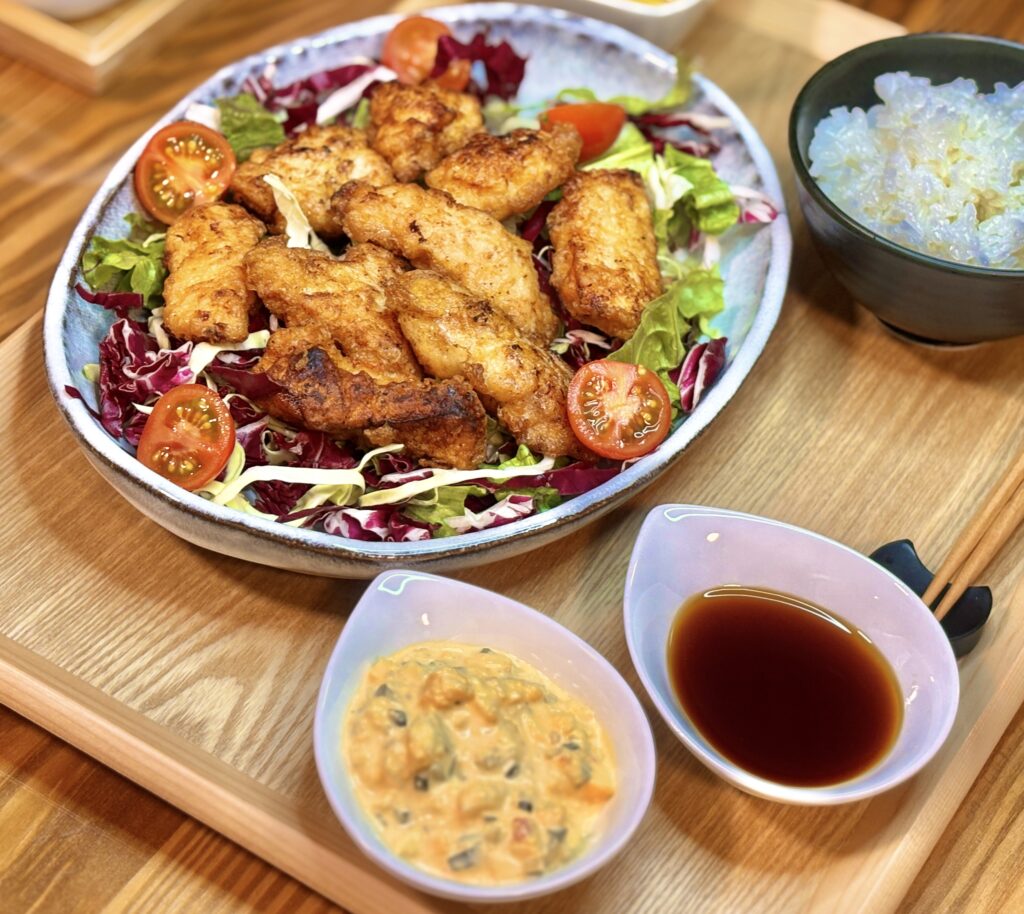 斜め上のアングルから撮影された、ボリューム満点のチキン南蛮定食。彩り豊かな野菜の上に一口サイズのチキン南蛮が盛り付けられ、手前にはたっぷりのタルタルソースと甘酢ダレ、奥にはマグロとアボカドの小鉢と炊きたてのご飯が並ぶ、旦那様のこだわりが詰まった食卓の風景。