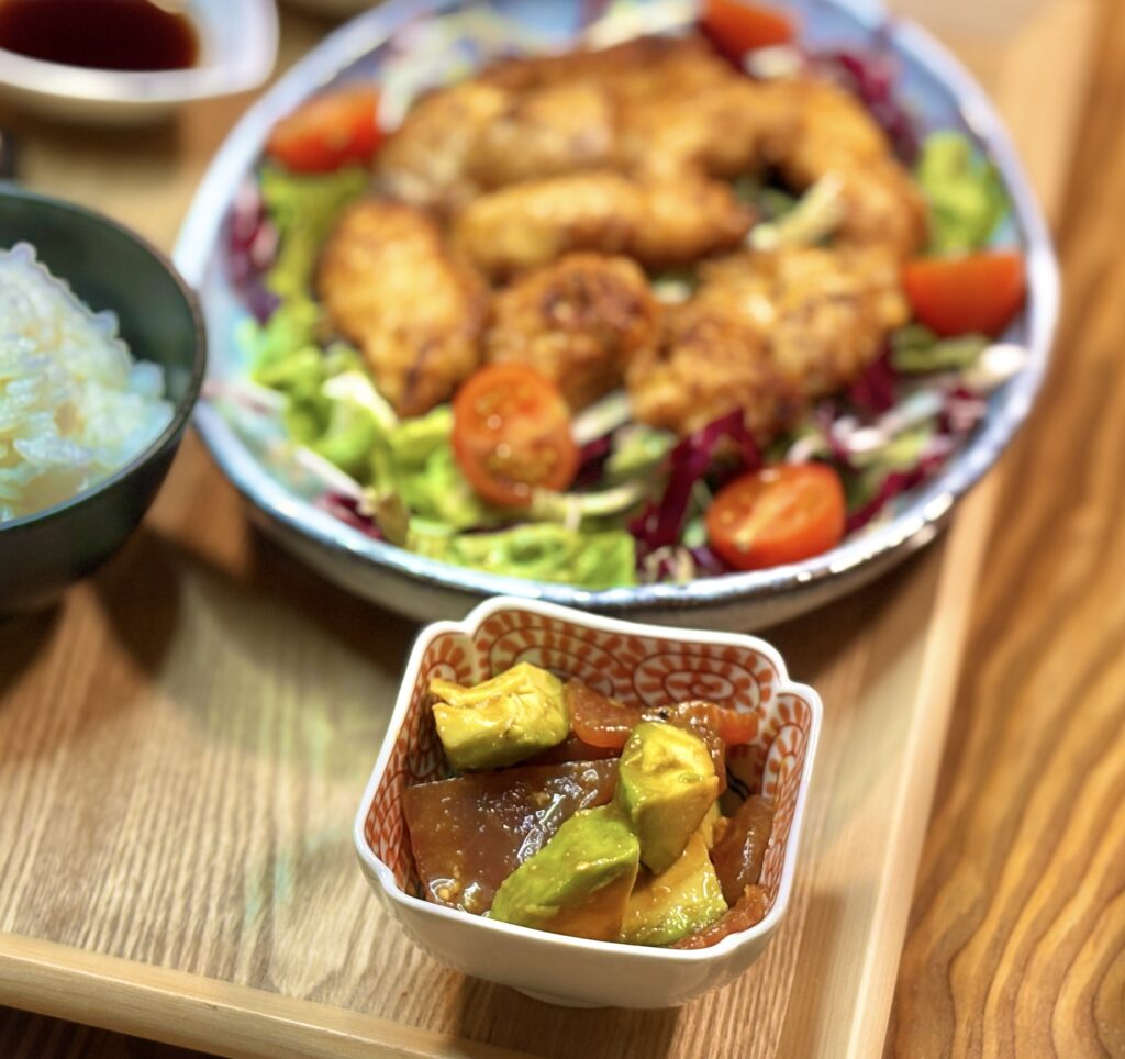 和柄の小鉢に盛り付けられた、マグロとアボカドのワサビ和え。明るい色味の木製トレーの上に置かれ、背景にはメイン料理のチキン南蛮がうっすらと見える、食欲をそそる彩り豊かな副菜のクローズアップ。