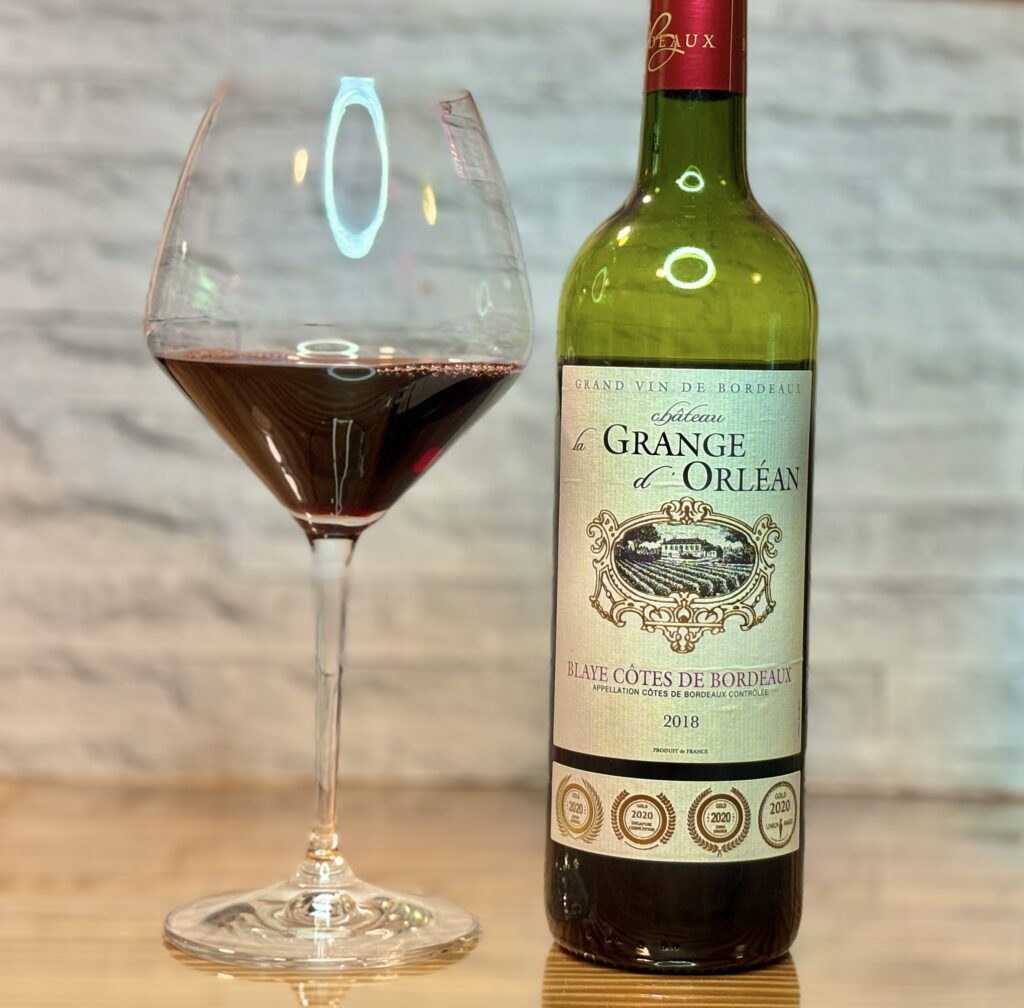 「4冠金賞」のメダルが輝くボルドー赤ワイン「シャトー・ラ・グランジュ・ドルレアン」のボトルと、なみなみと注がれたワイングラス。白煉瓦風の壁を背景に、反射するテーブルの上に美しく佇む、優雅な晩酌の風景。