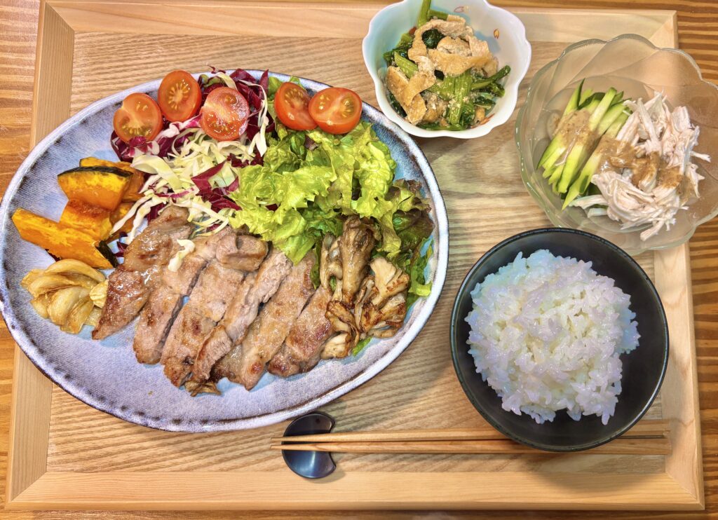 木製のトレイに並べられた本日の旦那キッチン定食全体図。メインのポークソテーに加え、白ごはん、棒棒鶏、ほうれん草の煮浸しがバランスよく配置されている
