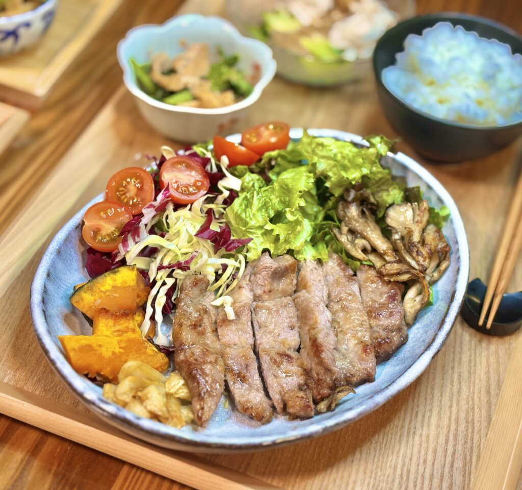 青い陶器の皿に盛り付けられた、ジューシーなガーリックポークソテー。付け合わせにはグリルの焼き目がついた舞茸、ホクホクのカボチャ、新鮮なレタスとトマトのサラダが添えられている
