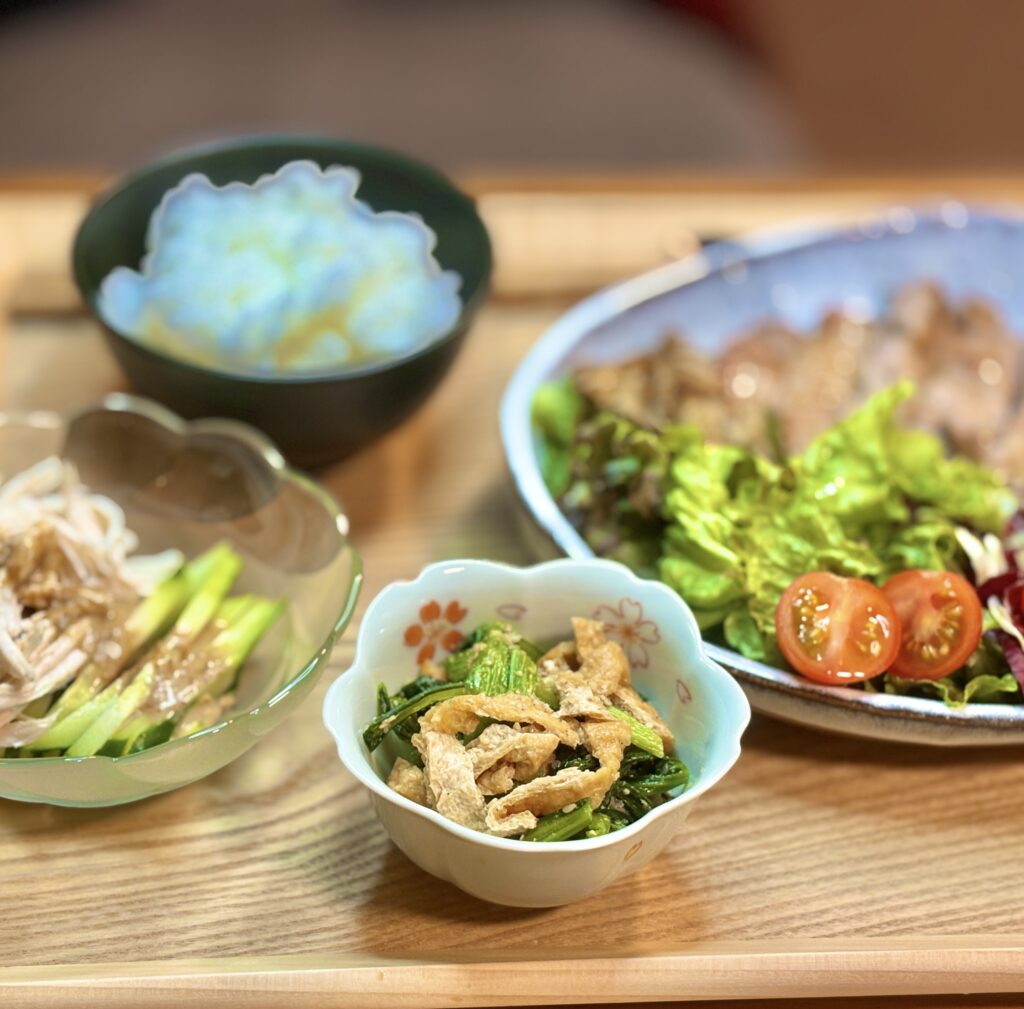 副菜2品のクローズアップ。花柄の小鉢に入った稲荷揚げとほうれん草の煮浸しと、ガラスの器に涼しげに盛られたしっとり鶏肉とキュウリの棒棒鶏
