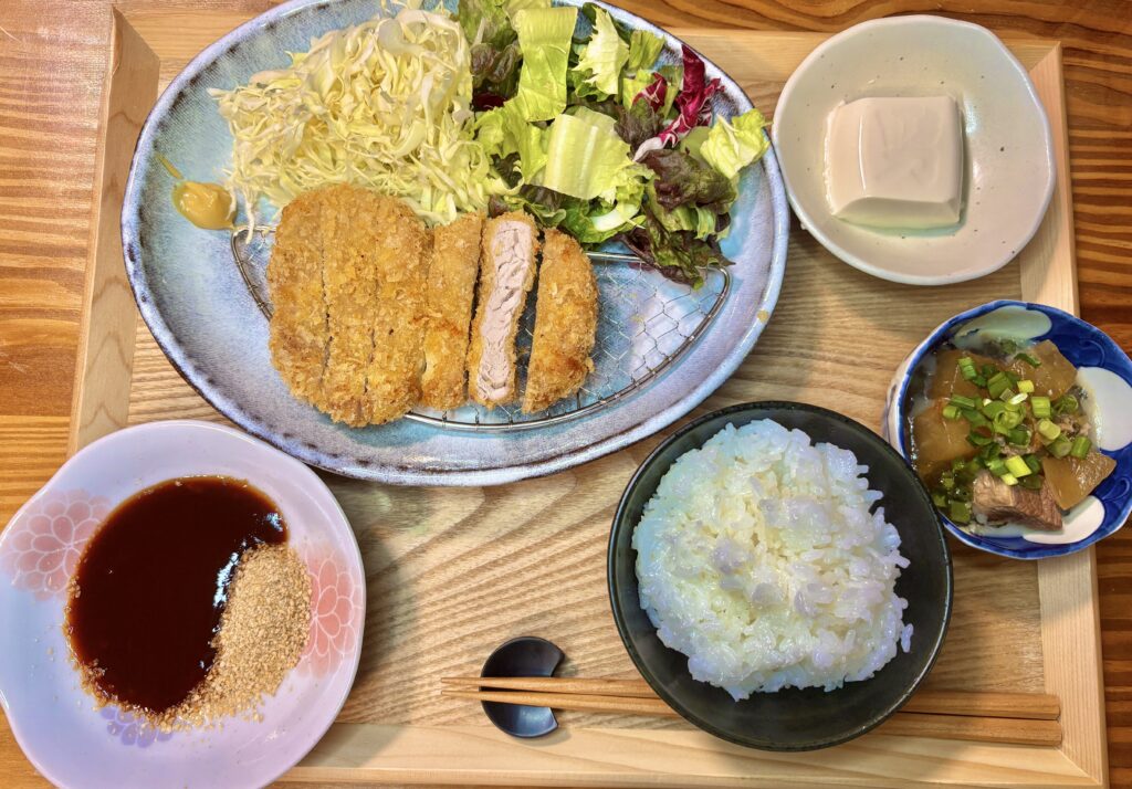 木製のトレーに並べられたヒレカツ定食の全体を真上から撮影した写真。中央の青い皿にはカットされたヒレカツと千切りキャベツ、レタス。左手前にはすりごま入りソース、右手前にはご飯、右奥には大根と牛肉の煮物、冷奴の小鉢が配置されている。