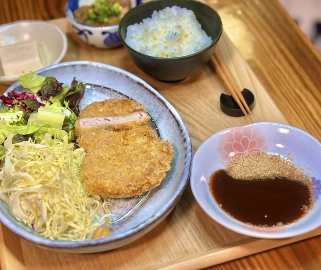 斜め上から撮影されたヒレカツ定食の写真。手前右側のピンク色の小皿に入った、たっぷりのすりごまとソースが強調されている。その奥にサクサクのヒレカツとキャベツが盛られたメインの皿がある。