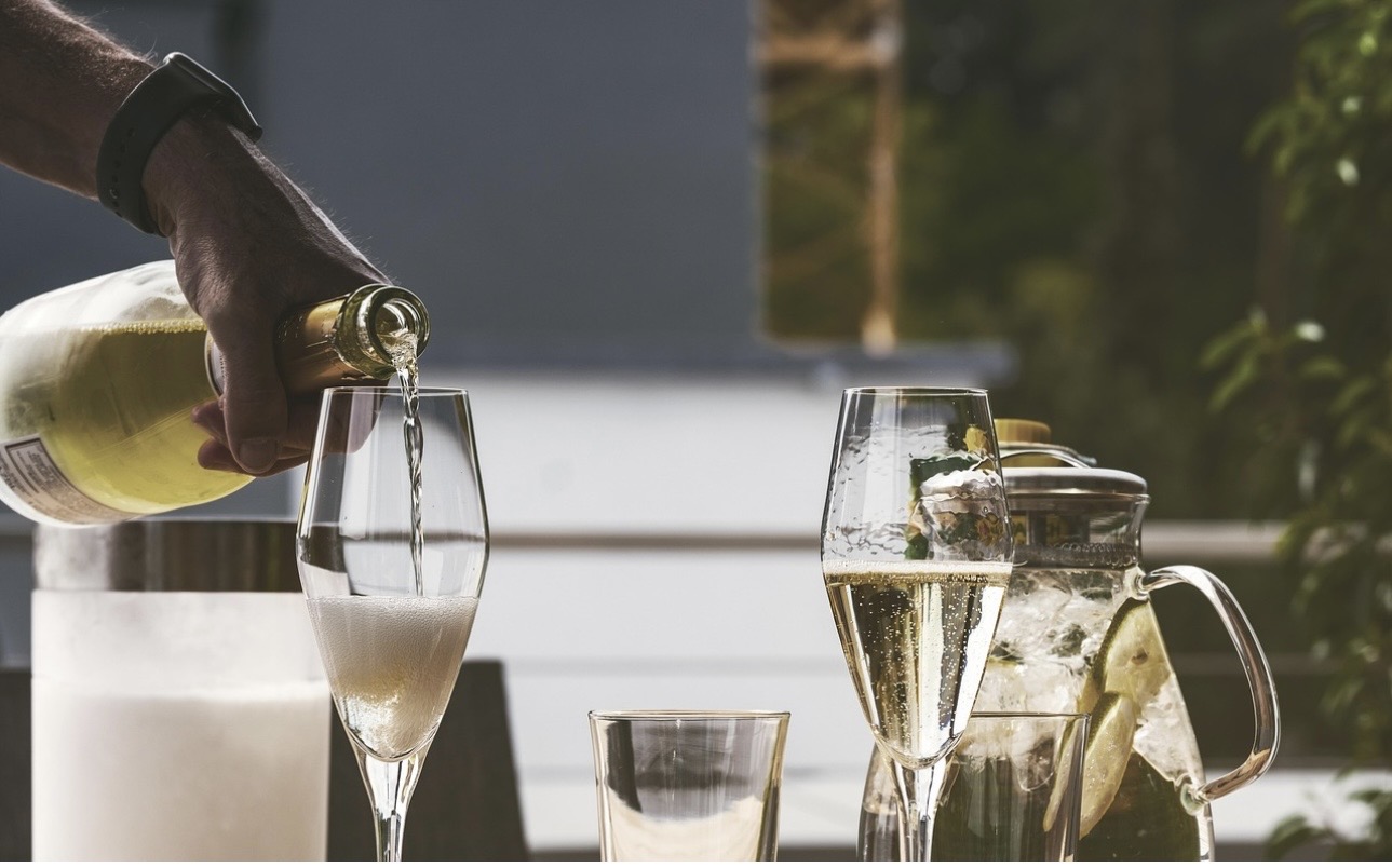 屋外のテーブルで、ボトルからシャンパングラスにスパークリングワインを注いでいる様子。隣にはライムと氷がたっぷり入ったピッチャーが置かれている。"