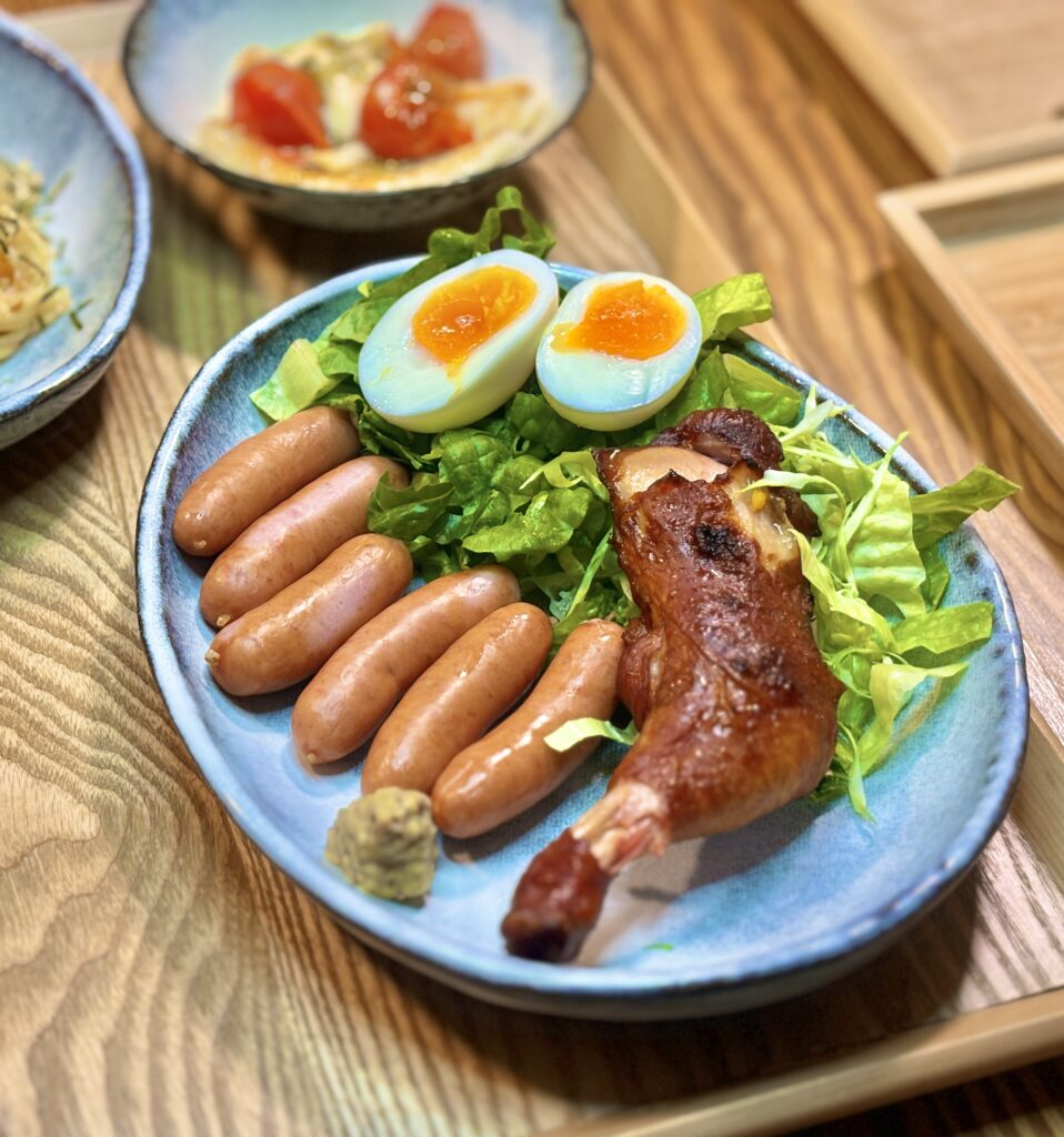 こんがり焼けた骨付きローストチキンとボイルしたシャウエッセンのアップ。黄身が濃厚な半熟卵が食欲をそそる、晩酌にぴったりのメインディッシュ