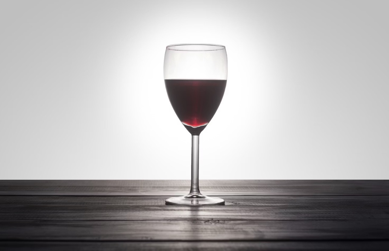 逆光に照らされた一杯の赤ワイン。暗い木製テーブルの上で、ワインの深い赤色が鮮やかに透き通り、グラスのシルエットが美しく浮かび上がっている様子。