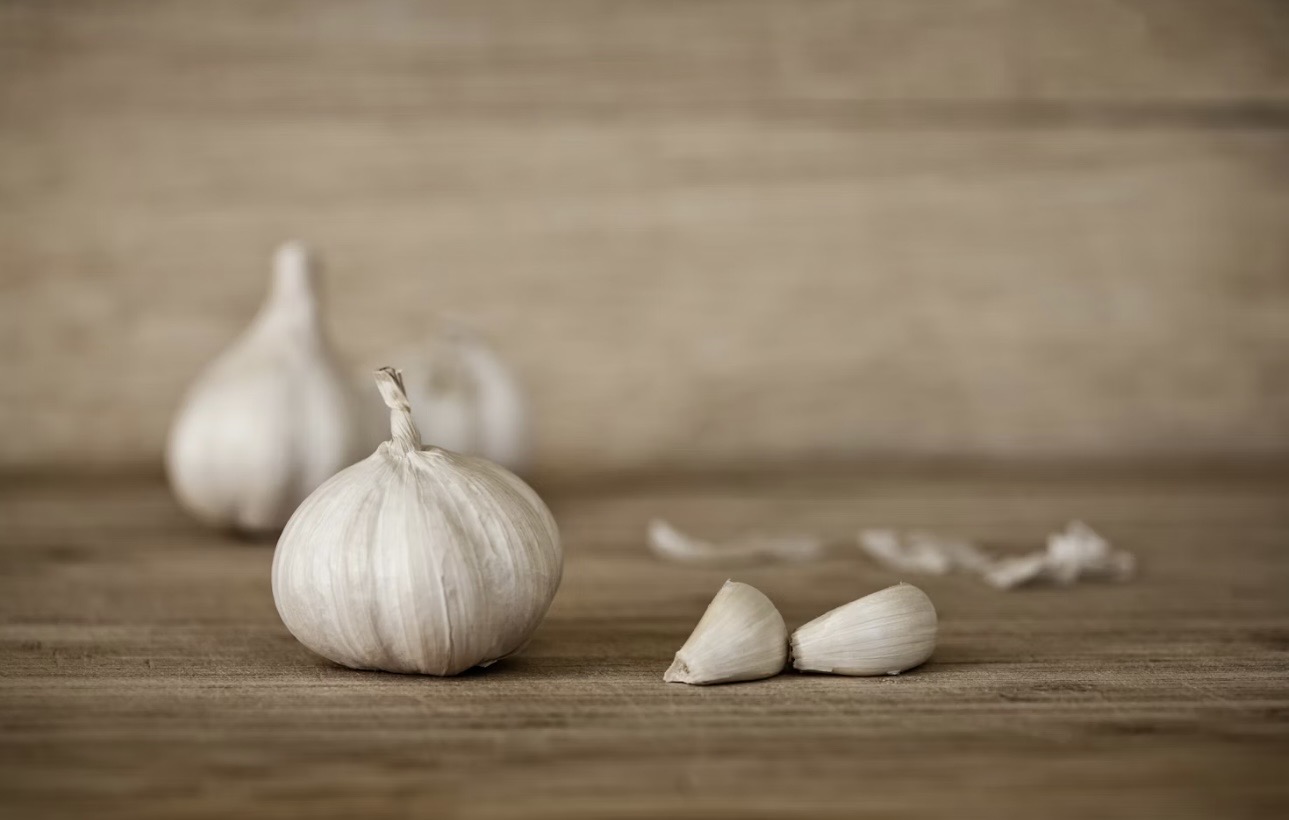 木目の美しいテーブルに置かれた白いニンニク一玉と、その横に並ぶ皮をむいた二片のクローズアップ。背景はボケており、新鮮でナチュラルな素材の良さが伝わるイメージ