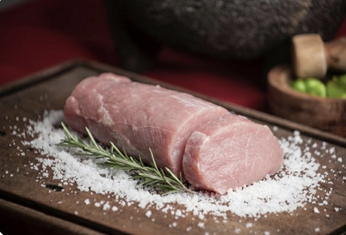 木製のカッティングボードの上に置かれた、生の豚ヒレ肉のブロック。周囲には粗塩が散りばめられ、ローズマリーの小枝が添えられている。背景にはすり鉢とライムが入った木のボウルがぼんやりと見える。