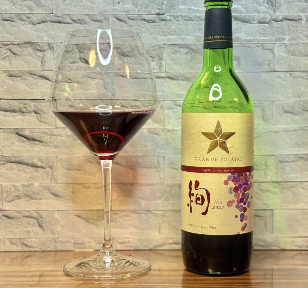 赤ワイングラスと日本ワイン「グランポレール 甲州 綾 2023」を並べた一枚。石調の壁を背景に、落ち着いた雰囲気で楽しむおうちビストロのワインシーン。
