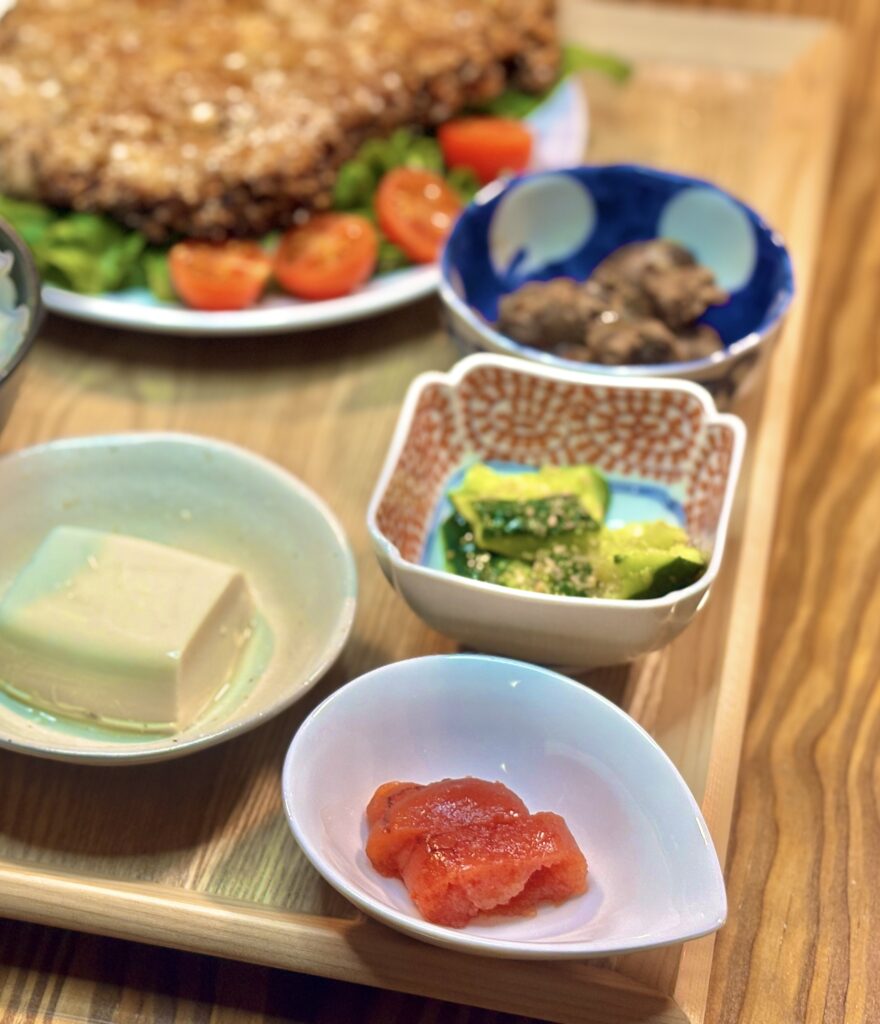 手前中央にある白いしずく型の小鉢に盛られた明太子と、その奥に並ぶ赤い柄の器の叩ききゅうり、青い小鉢のレバーの甘露煮、冷奴の食卓写真
