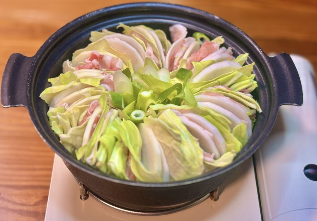 黒い土鍋に、豚バラ肉・大根・キャベツを放射状に重ねた生姜アゴだしのミルフィーユ鍋が盛られている。中央には刻みねぎが添えられ、コンロの上で火にかかっている調理中の様子。