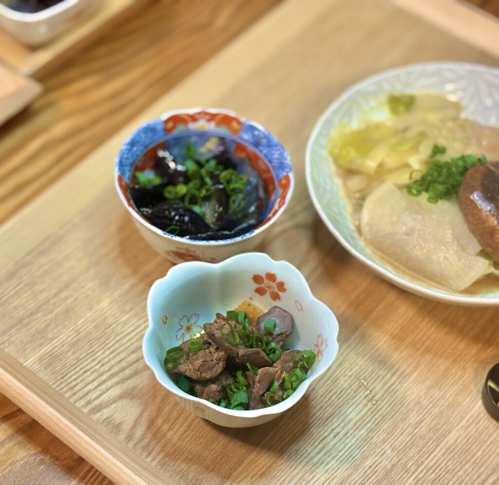 手前に鳥レバーの甘露煮、奥に揚げ出し茄子、右側にミルフィーユ鍋の取り分け皿が配置されている。木目のテーブルと器の色合いが温かみを演出している。